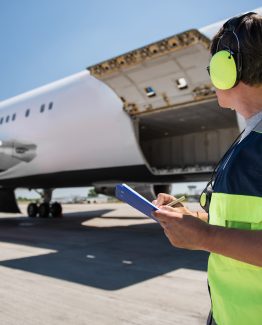Aircraft ground handling. Man in headphones observing airplane with open cargo door and intending to fill out documents. Copy space in left side